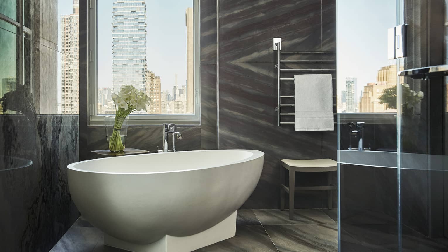 Free-standing white bathtub under tall window, modern grey tile bathroom