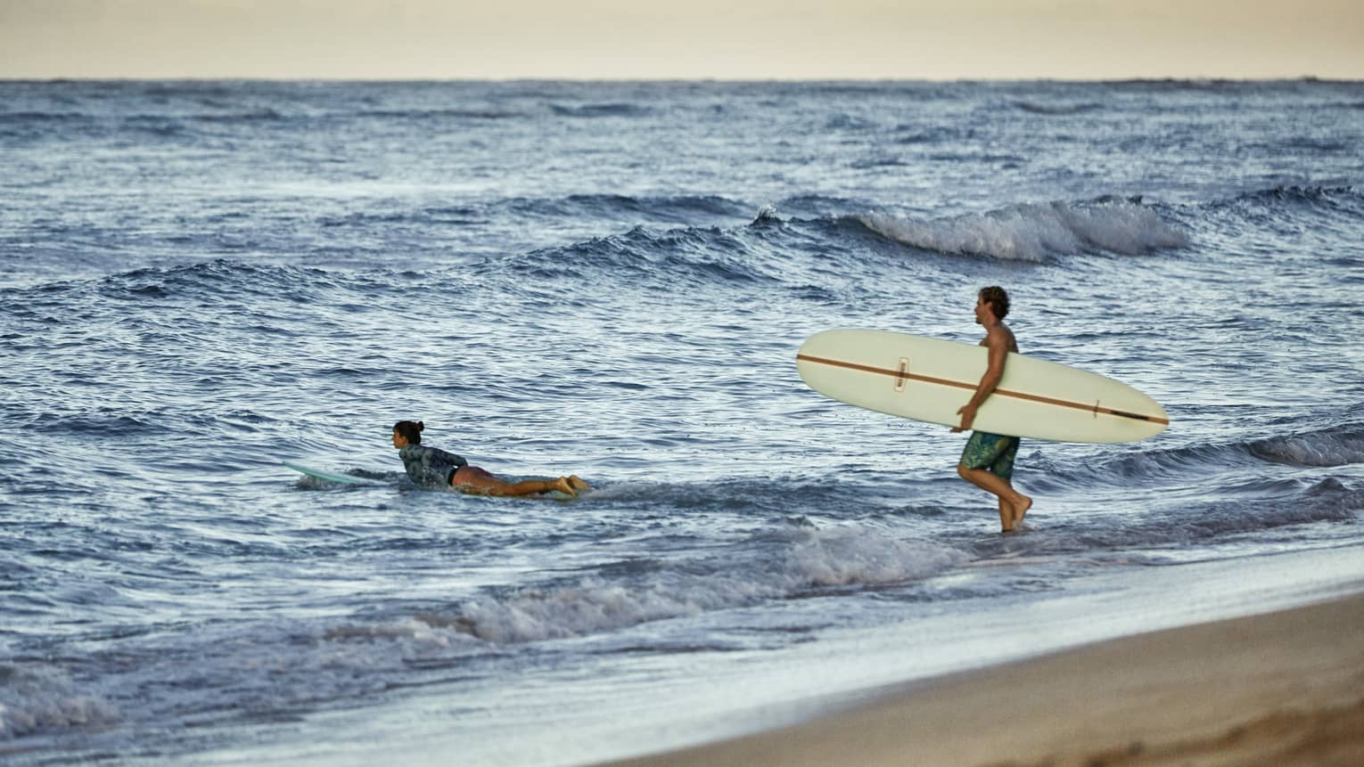 Under a golden-yellow sky at the beach, a surfer paddles their board into the waves as another wades in carrying a board.