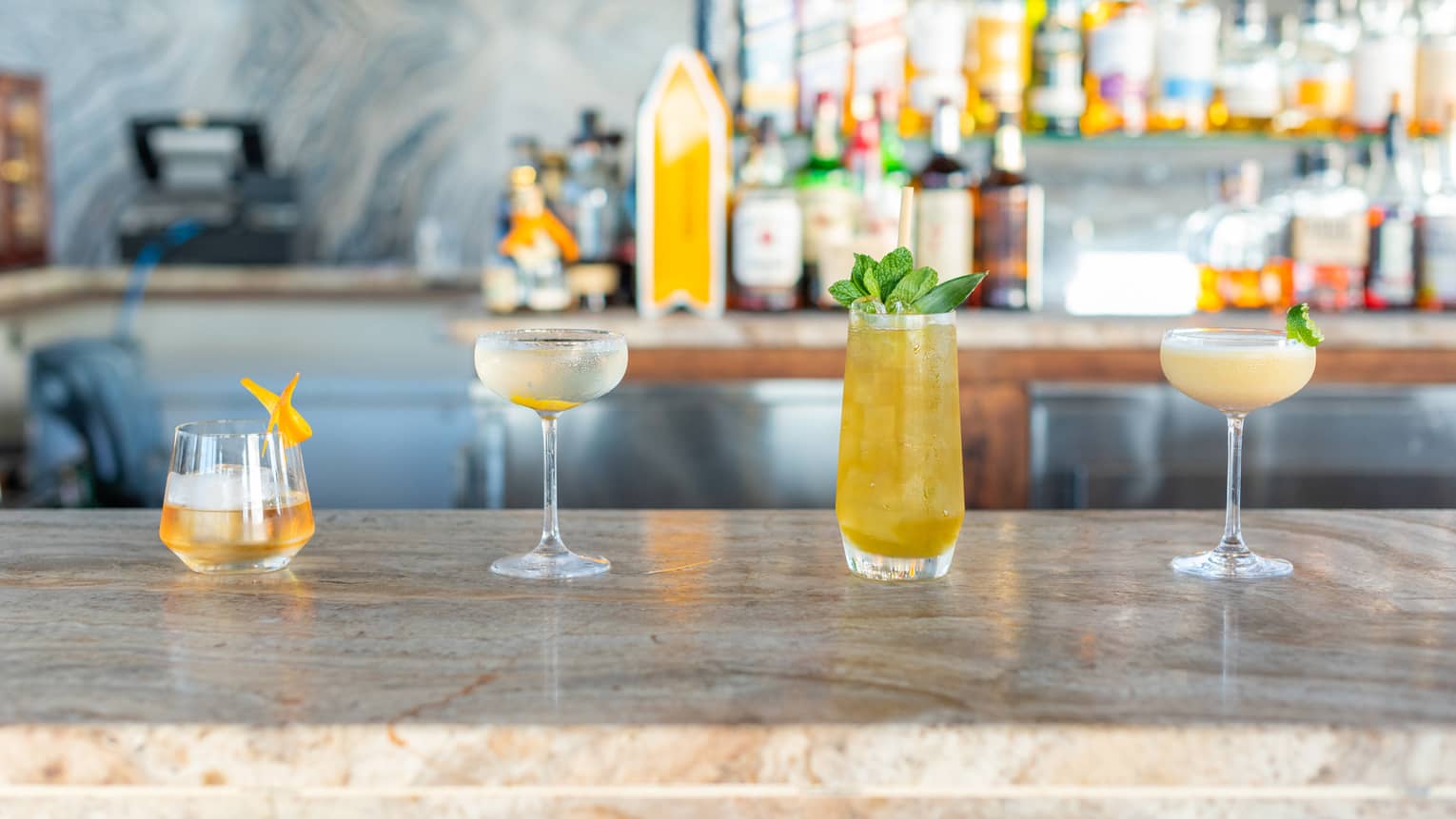 Four cocktails on a bar counter with bottles behind them.