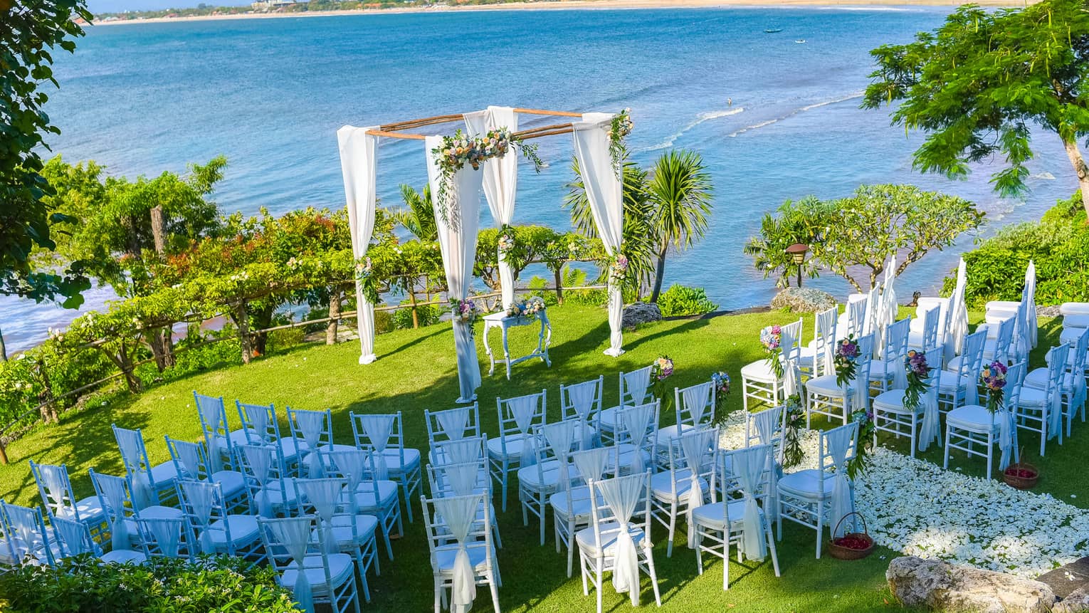 Aerial view of rows of white chairs facing wedding altar on lawn by ocean