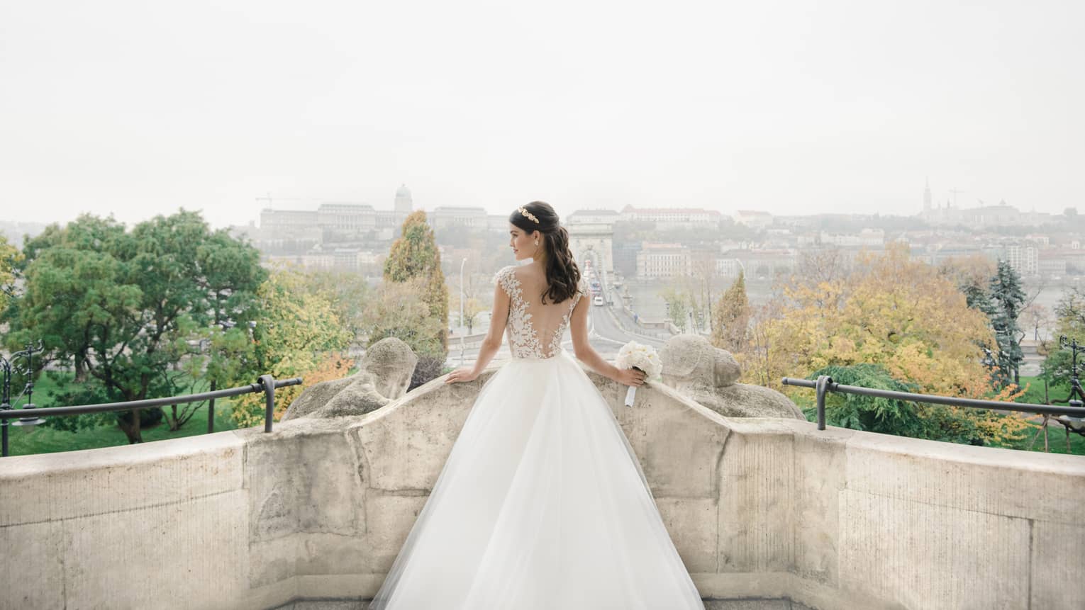 Rear view of bride on stone terrace, looking to the side and holding white bouquet, Budapest view