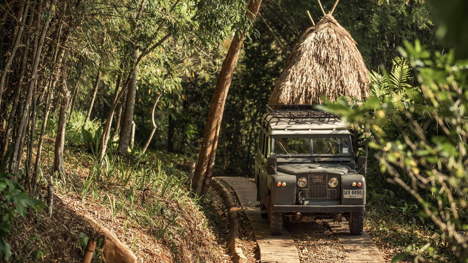 Jeep driving down path in forest past thatched-roof hut