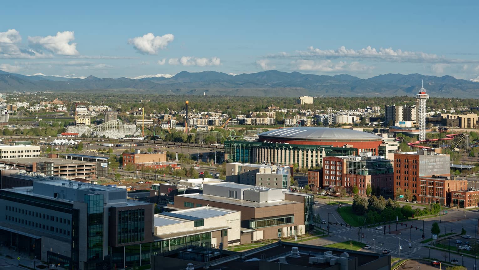 Aerial city view featuring an amusement park, a large round building, a TV tower, mountains in the distance under blue sky.