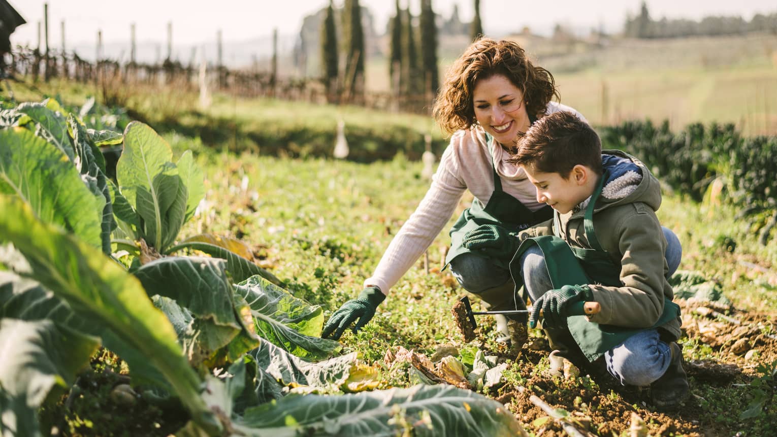 Woman and boy wearing aprons, gloves kneel in soil near large lettuce leaves in vegetable garden patch