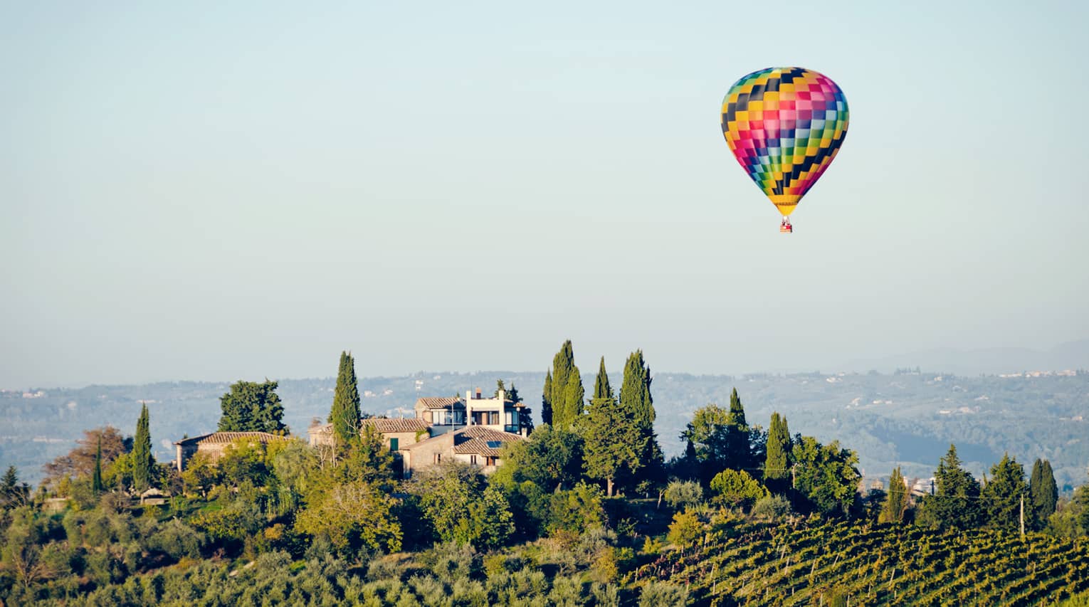 Colourful hot air balloon floating above a landscape with trees and houses, set against a clear blue sky.		