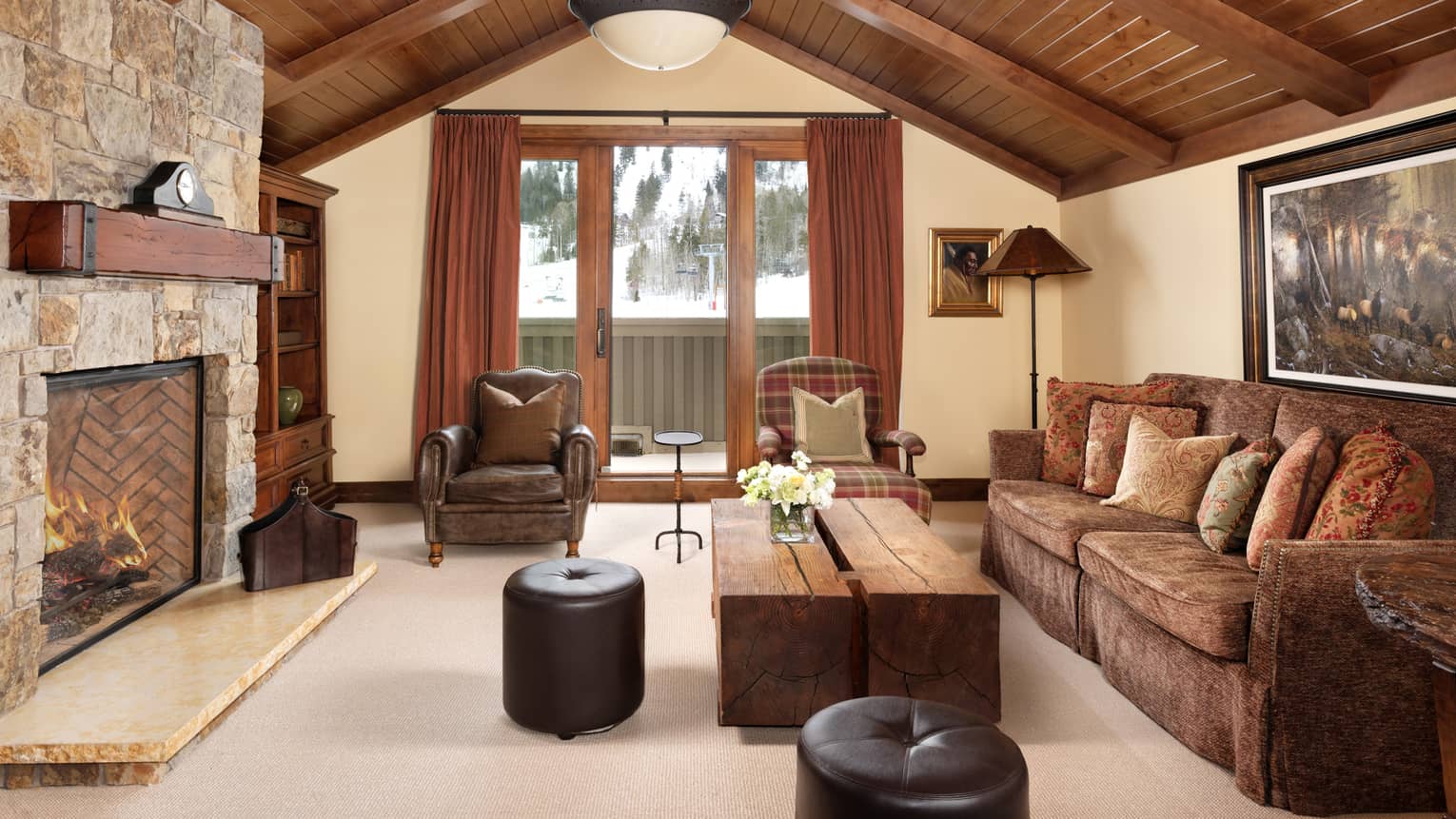 Living room with vaulted wood ceiling, stone fireplace, wood coffee table, brown sofa, leather armchair and ottomans, and plaid accent armchair