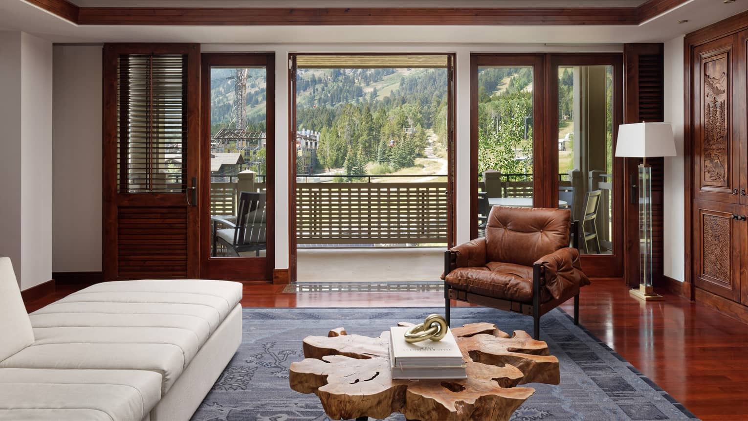 A hotel room with a white sofa, brown chair, wood coffee table and large window looking outside at hills and trees.