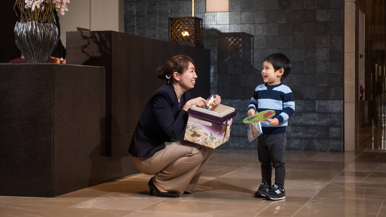 Woman with toy box kneels in front of smiling young boy holding fan