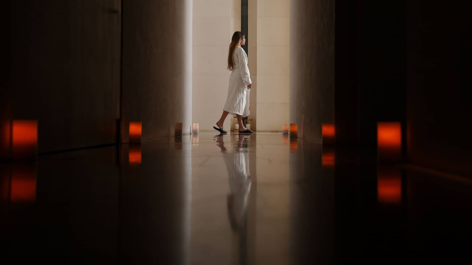Person with long dark hair wearing a white spa robe walks through a dark hallway lit by eight dim red lanterns