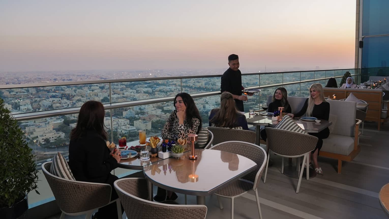People dining at restaurant tables set along the edge of an outdoor terrace overlooking a city skyline below