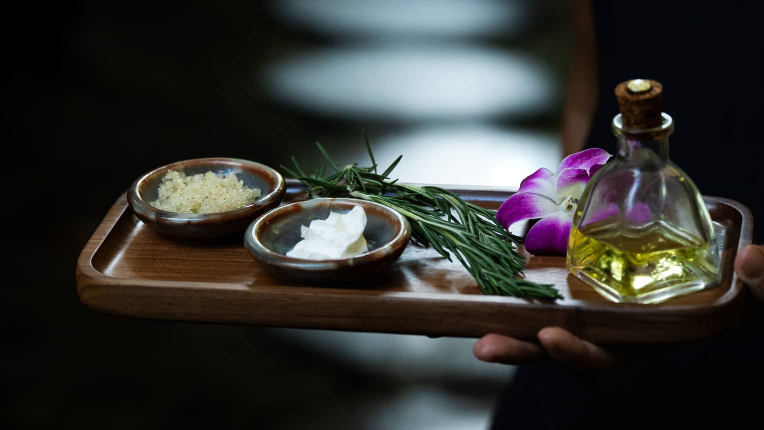 Hand holds a wooden rectangular tray with spa ingredients