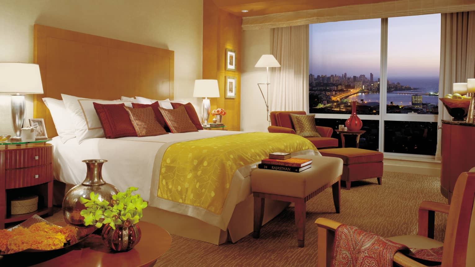 Deluxe Sea-View Room bed with yellow silk blanket, books on bench, chaise by window, sunset