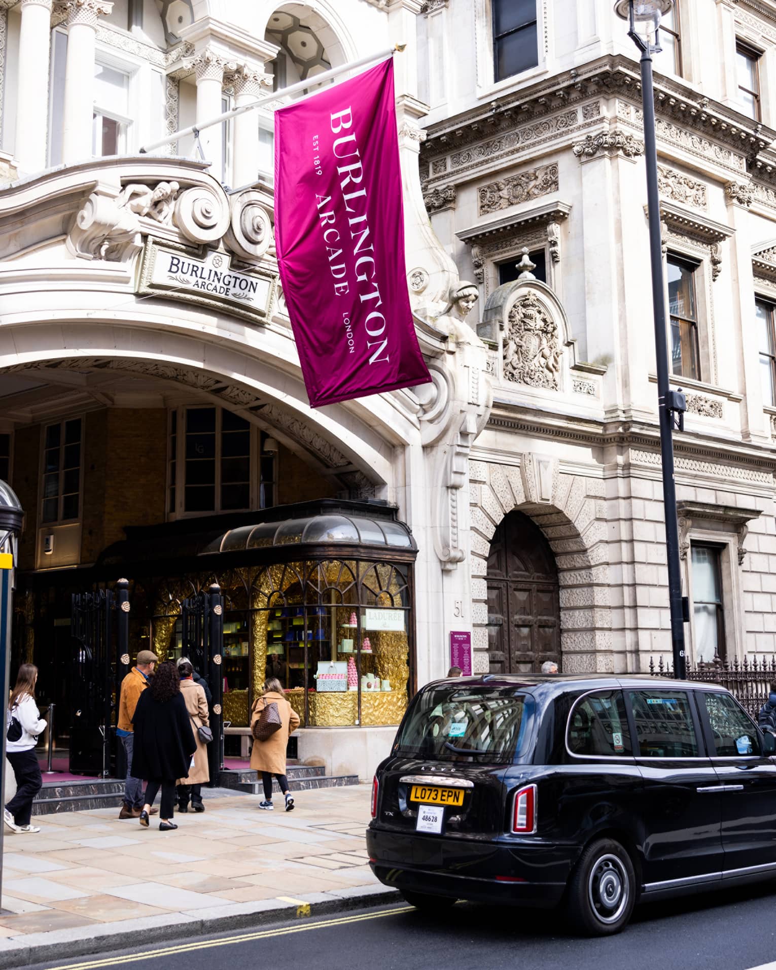 London-style black cab parked in front of a high-end pastry shop in a building with a Burlington Arcade sign and flag.