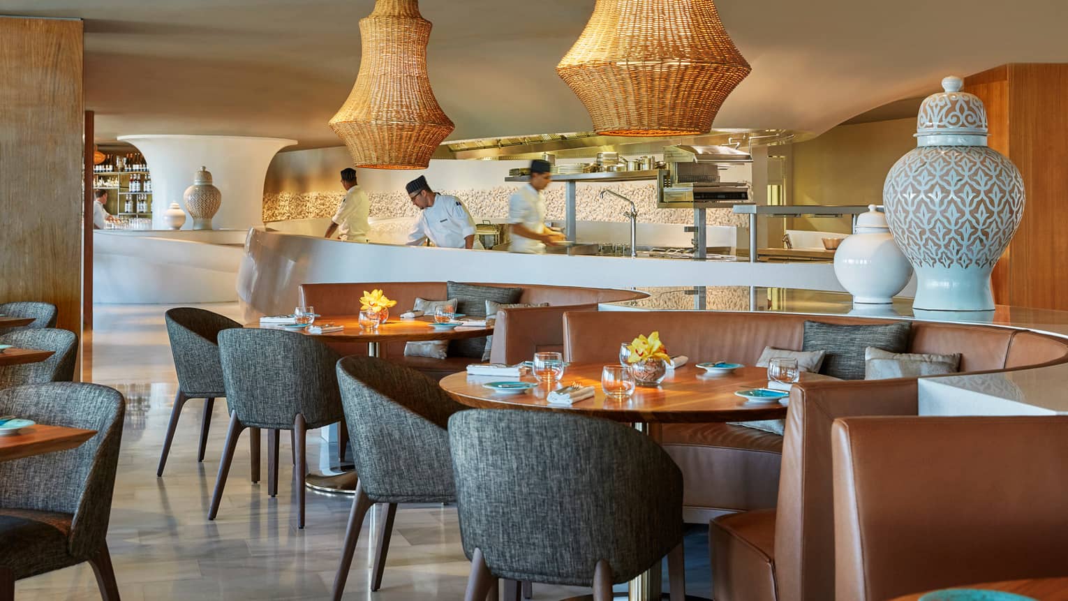 Large wicker lamps hang over mid-century style dining tables and chairs at Blue restaurant, chefs work in background