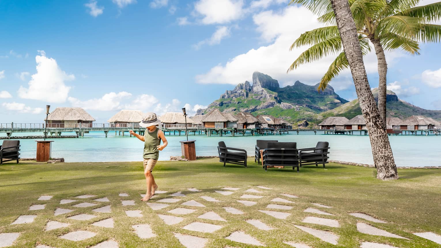 A child tiptoes on paving stones by a clear lagoon. Grass-roofed suites stand in the water in front of island mountains.