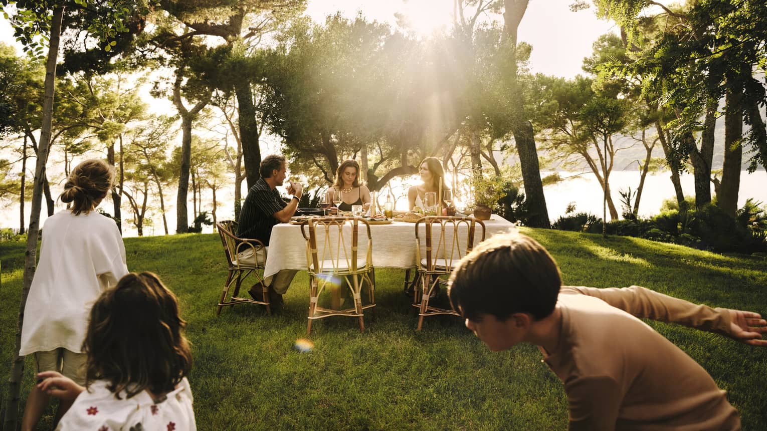 Children play in the foreground as adults dine at an ocean-view dinner table with the sunlight streaming through the trees.