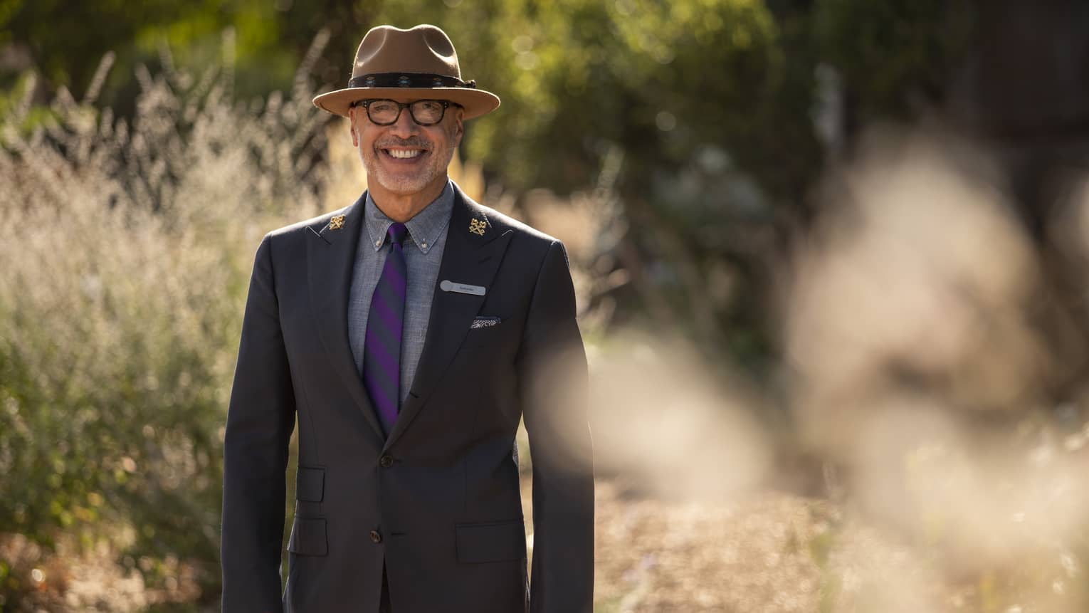 Four Seasons Chef Concierge Antonio Barrios smiles outdoors wearing a fedora and a dapper dark jacket with lapel pins.