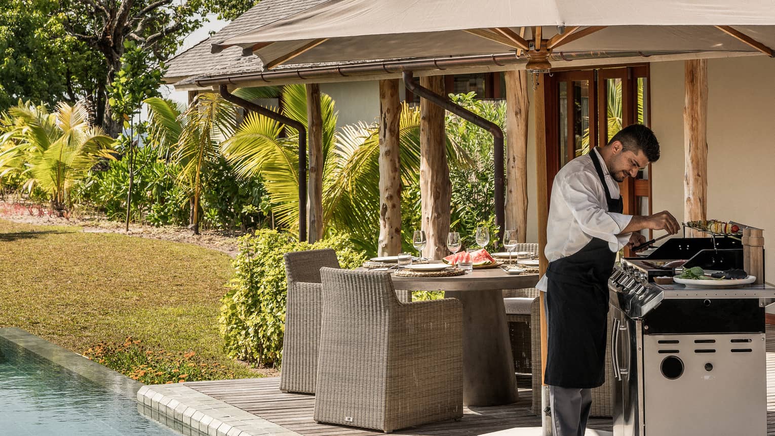 Chef wearing apron cooks at barbecue on deck by dining table, pool