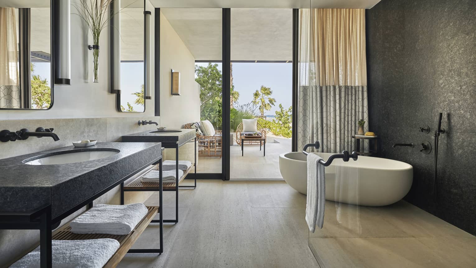 A bathroom with large sliding glass door leading to a sitting area, a bathtub and two sinks.