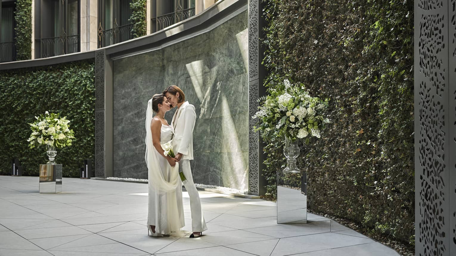 A couple in all white stands in a garden holding hands and looking into each others eyes.