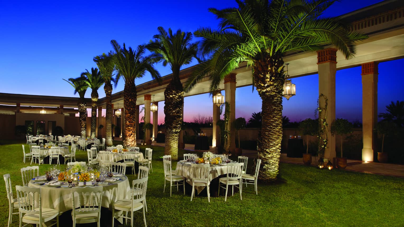 Le Jardin event lawn at night with small banquet tables, row of palm trees 