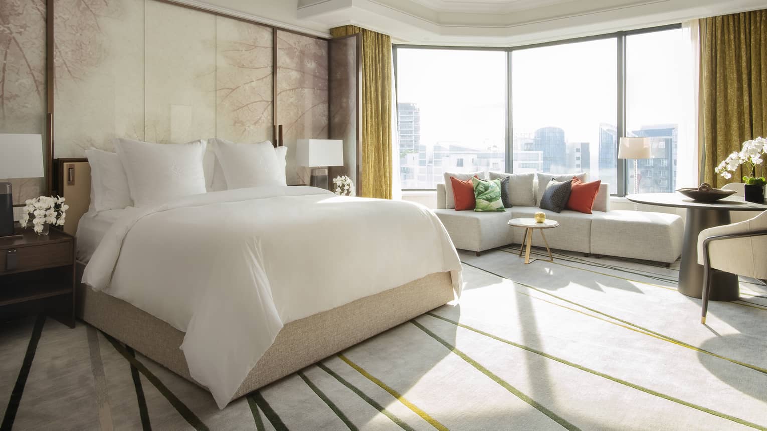 A spacious guest room featuring a large bed, seating area with colourful pillows and floor-to-ceiling windows with city views.