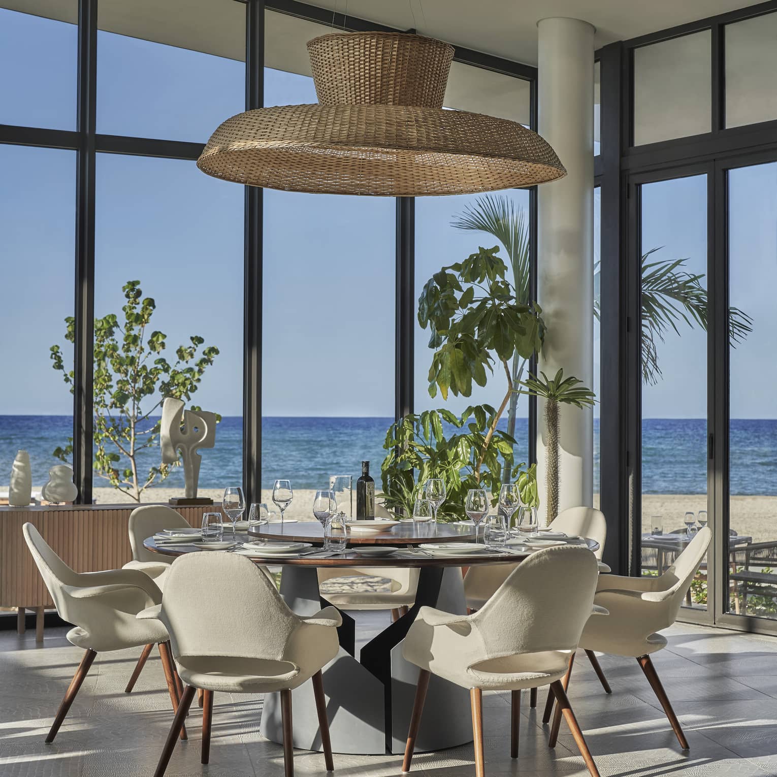 A round table with white leather chairs surrounding it and large floor to ceiling windows looking out at a beach.