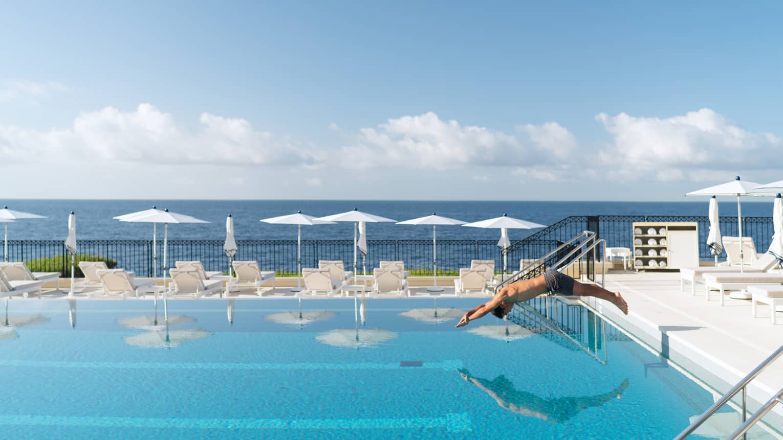 Man diving into blue Club Dauphin pool, white umbrellas and lounge chairs along edge, sea behind