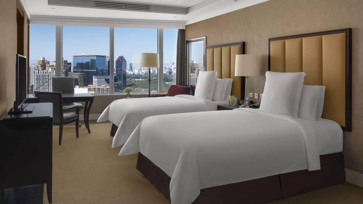 Hotel room with two single beds, tan headboards, dresser and TV, desk and two chairs