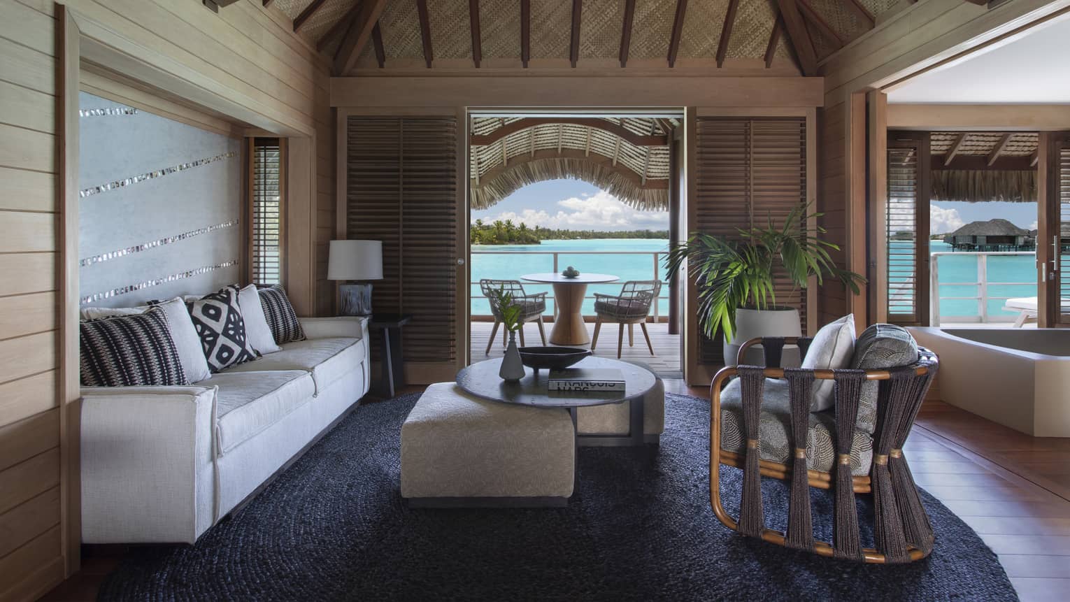 Overwater bungalow living area with neutral furniture, wood beams, tropical plants and a patio with a view of the turquoise lagoon. 