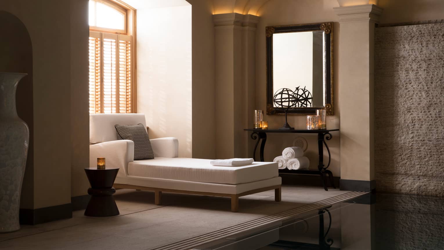 Long white chaise with accent pillow under sunny spa window with shutters, candles on shelves in room