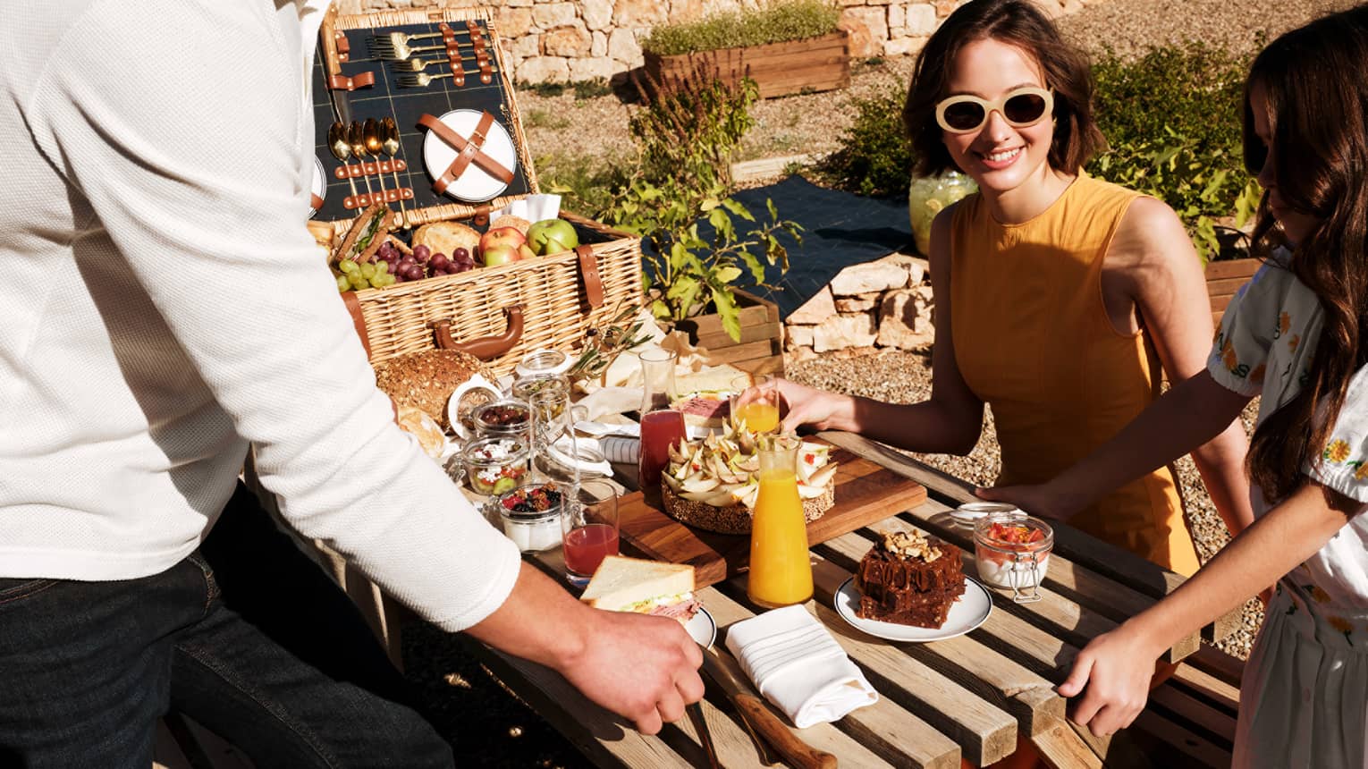 A young family enjoys a decadent picnic of sandwiches, fresh fruit, juice, tarts and brownies at a table bathed in sunshine.