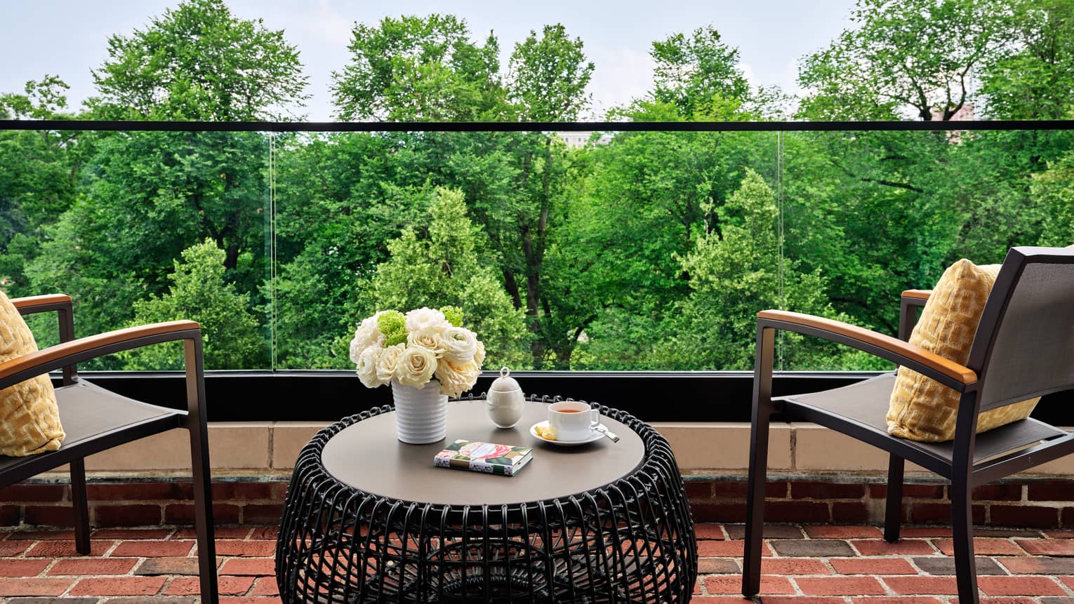 Private terrace of luxury hotel suite with two chairs, circular net-like table, and glass balcony looking out to gardens
