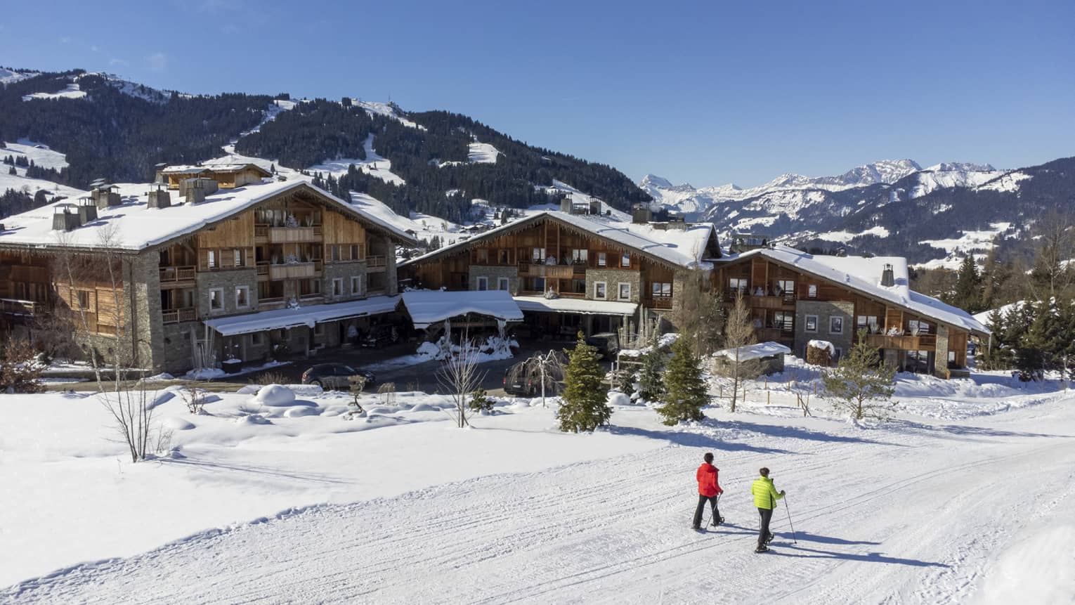 Two people show shoeing on snow-covered path in front of Four Seasons Hotel Megeve with Alps in backdrop