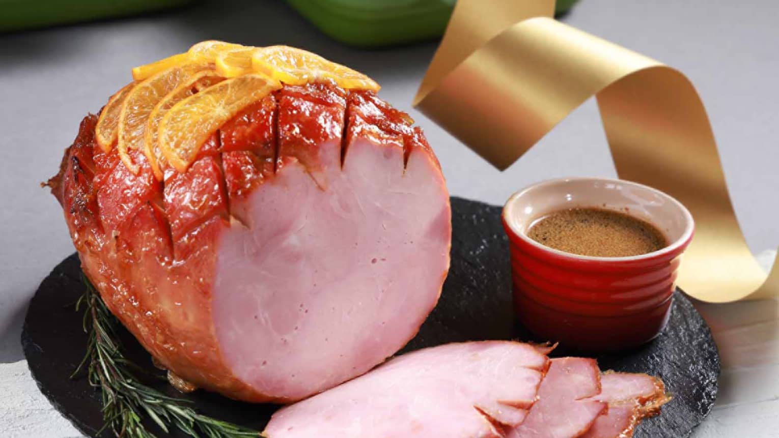 Glazed holiday ham topped with orange slices, served with a side of sauce in a red bowl on a black platter.