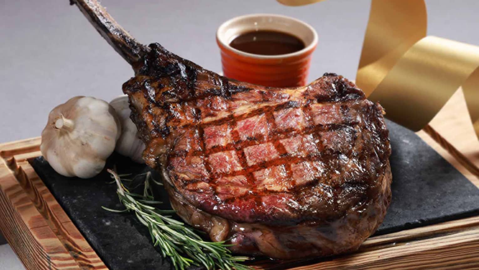 Grilled tomahawk steak with char marks, garnished with rosemary and garlic, served with a side of sauce in a red bowl.