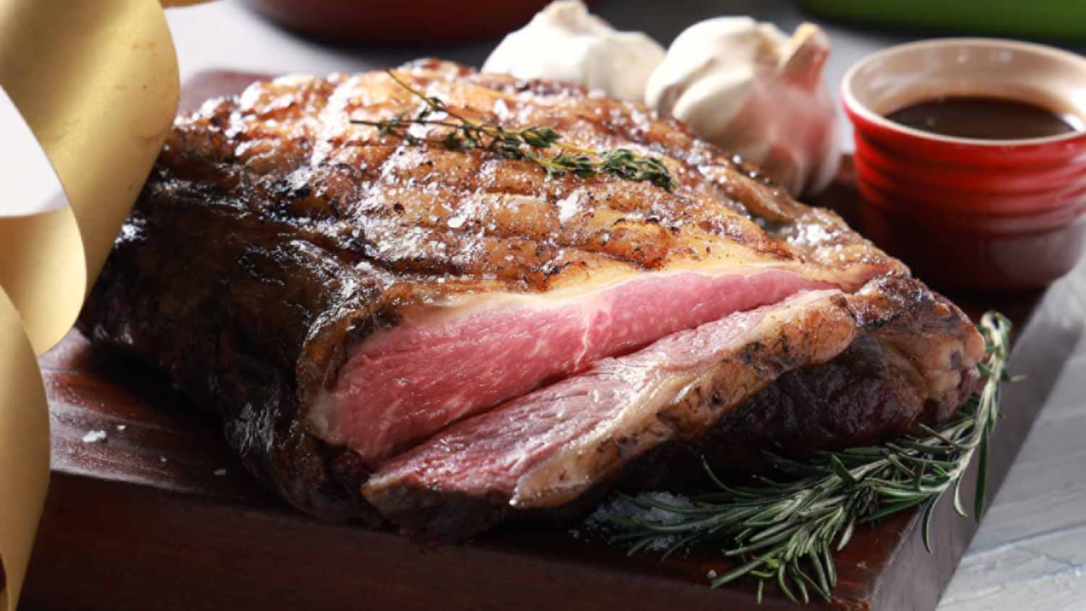 Medium-rare prime rib roast garnished with rosemary, served with garlic bulbs and a side of sauce in a red bowl.