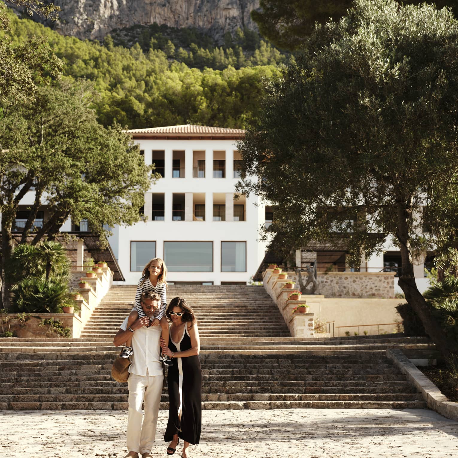 A family strolls in front of a grand stone staircase leading to an elegant white resort at the foot of a lush, treed cliff.