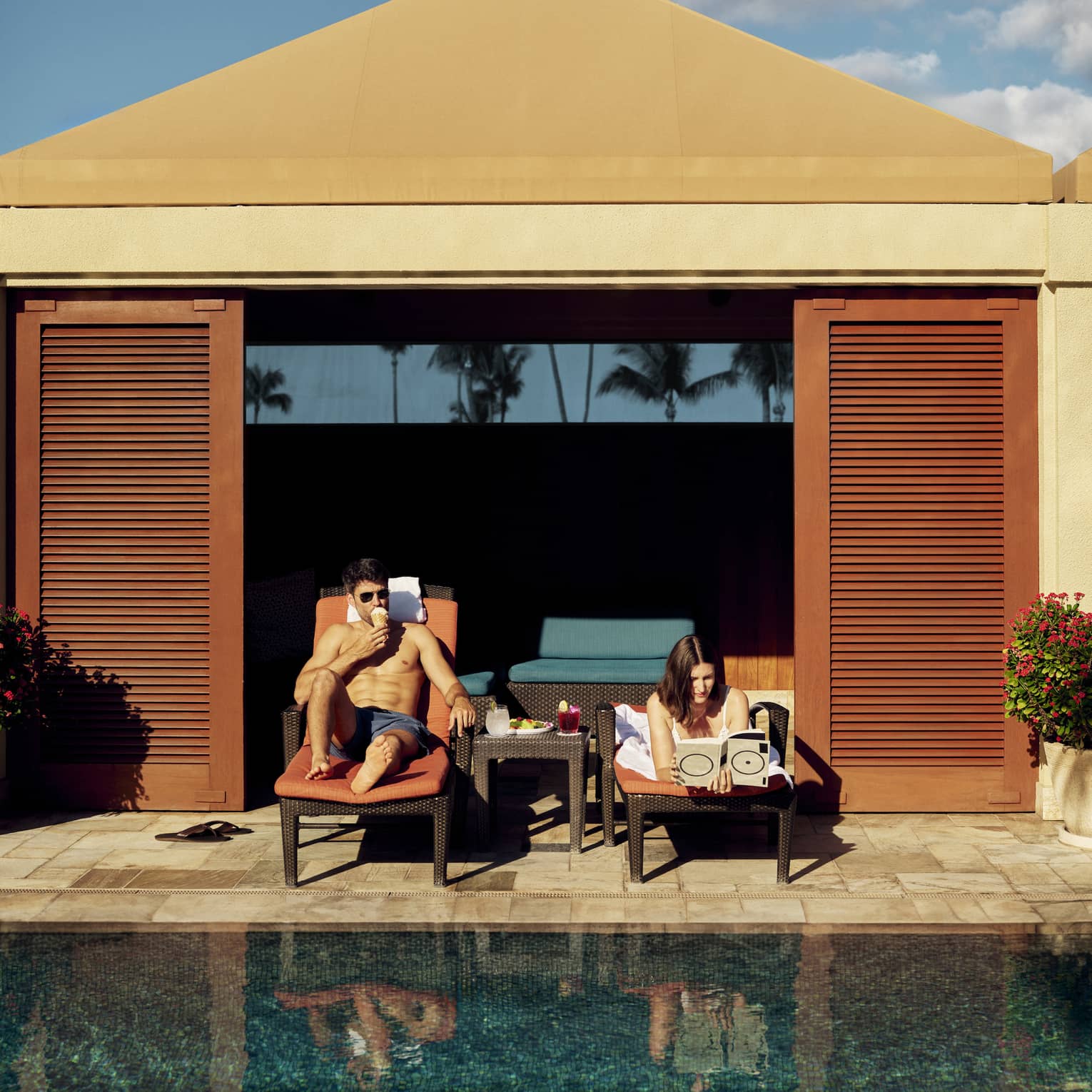Two people lounge on sun chairs in private poolside cabana