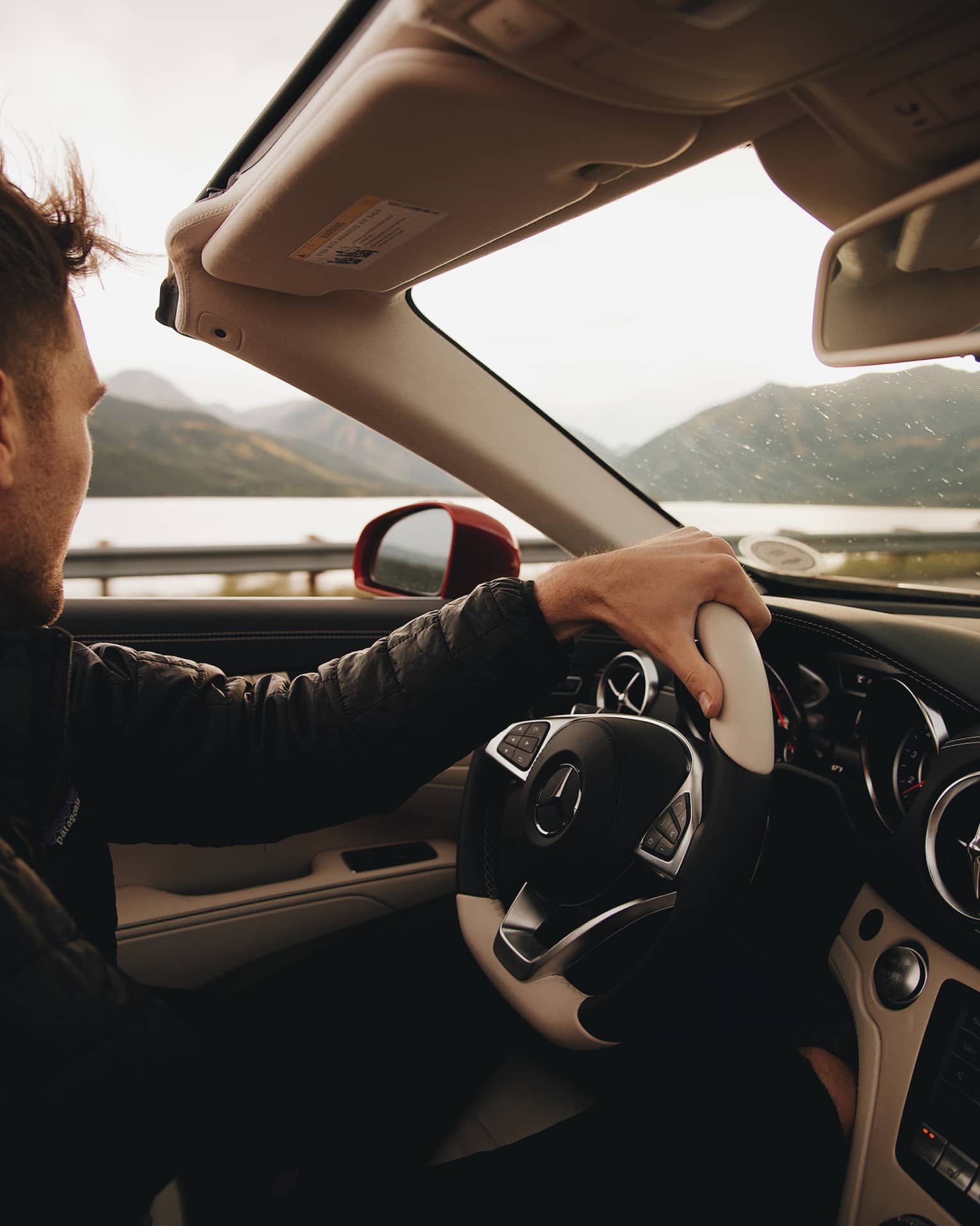 Passenger's view of driver gripping the wheel of a Mercedes convertible, looking away at a lake and distant mountains.