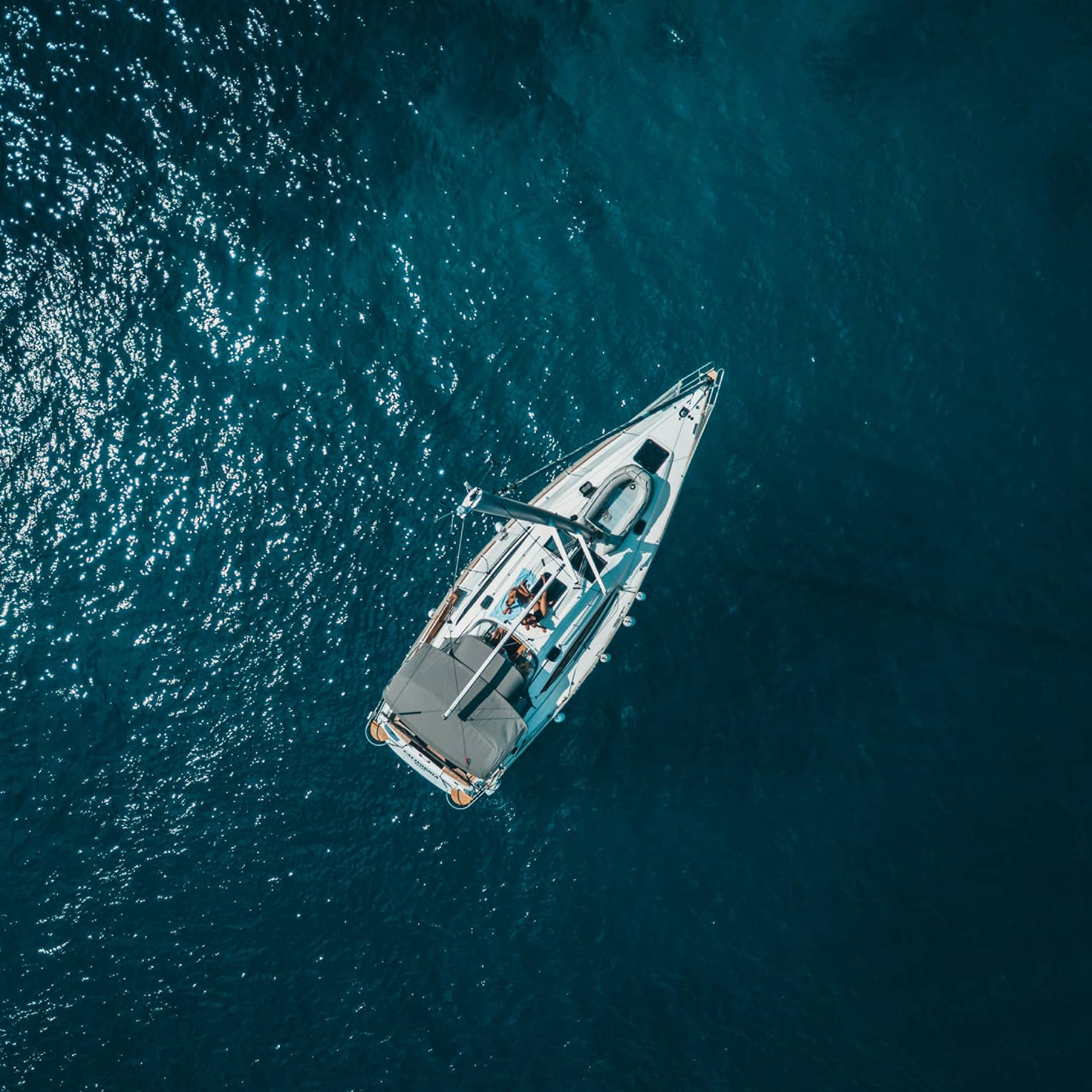 Distant aerial view of two guests sunbathing on a yacht surrounded by shimmering midnight-blue water.