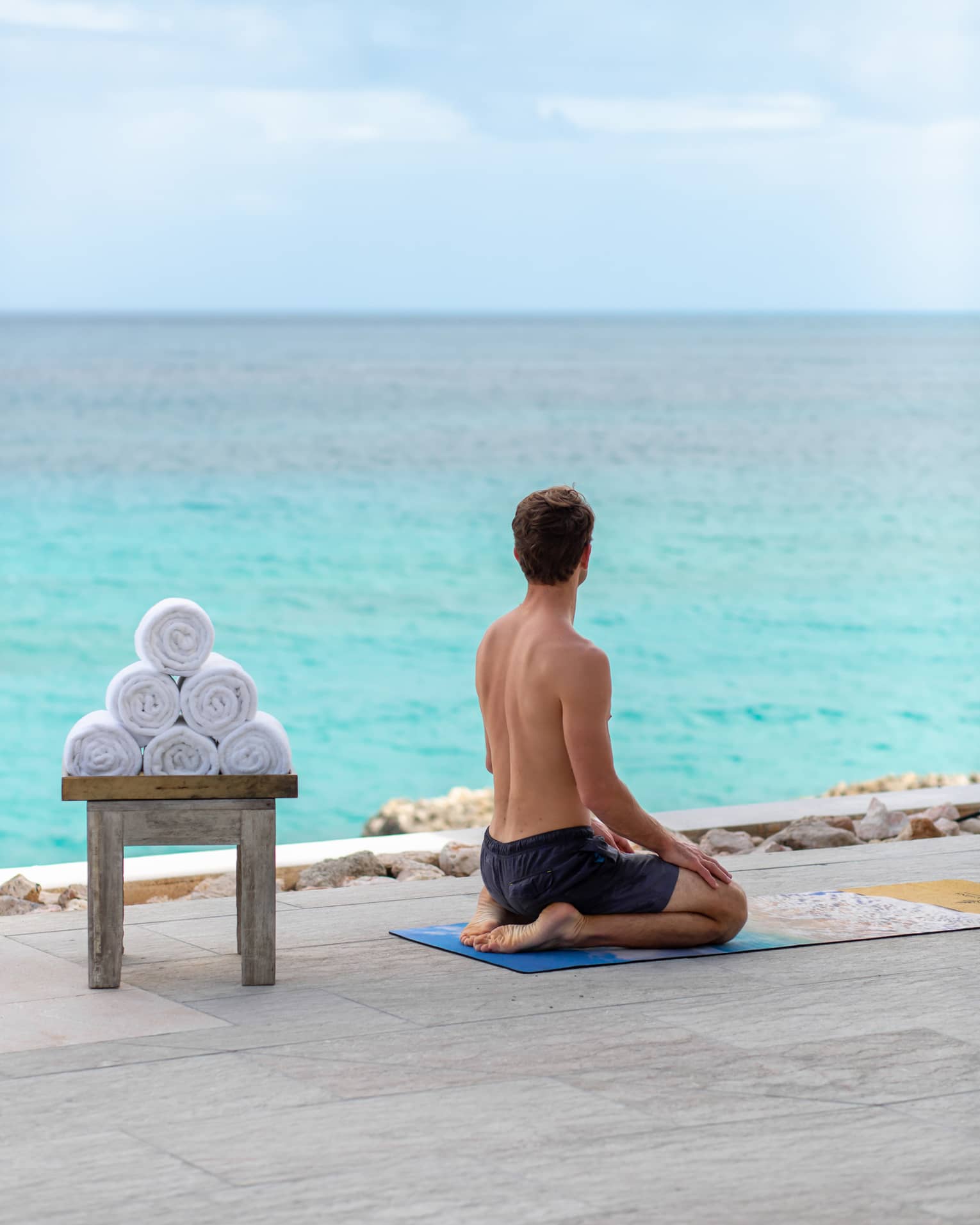 A man sitting on his legs with the ocean in front of him and towels on a bench behind him.
