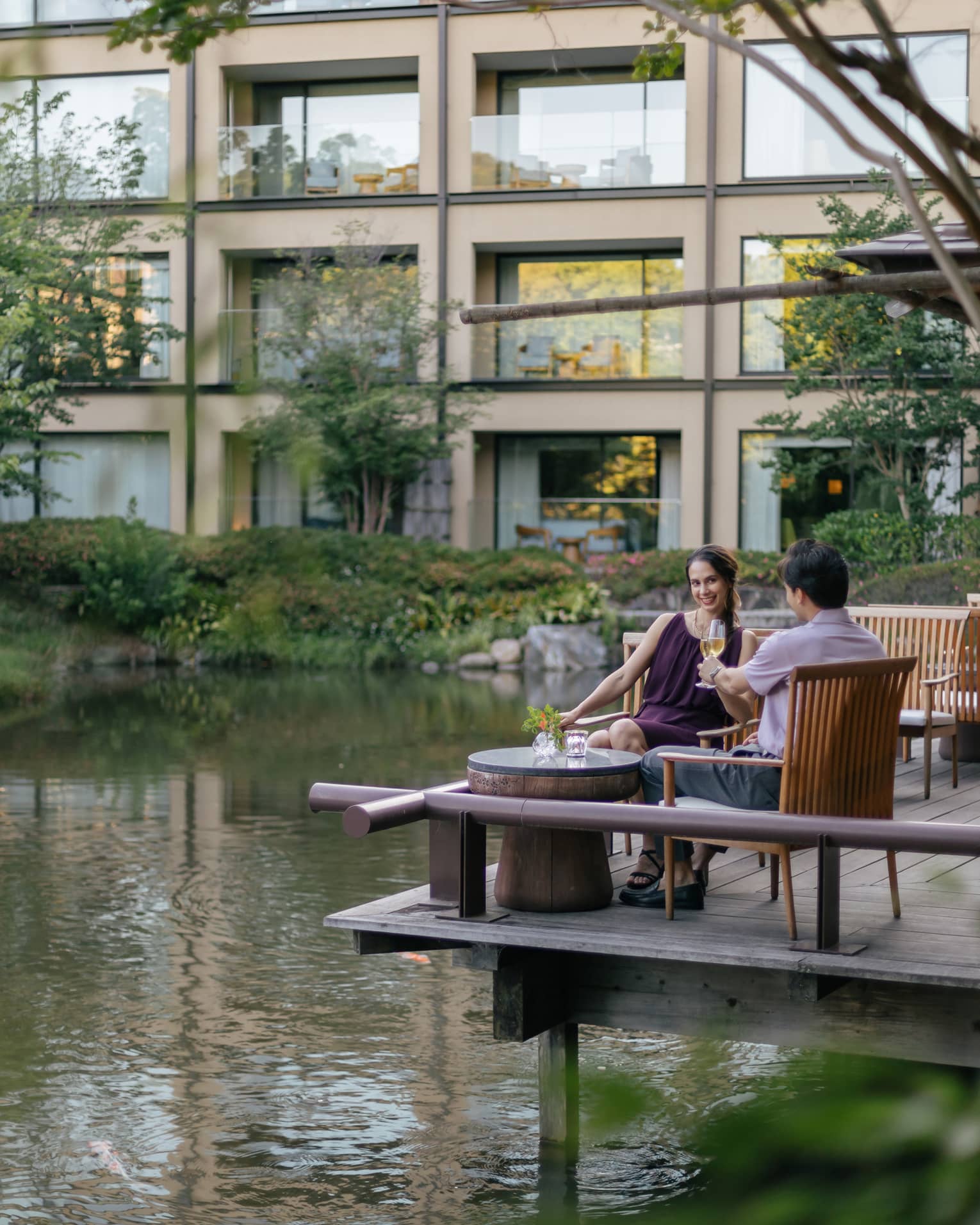 Smiling couple clink champagne flutes at a café table on a wood deck overlooking a rippling pond bordered by lush foliage.