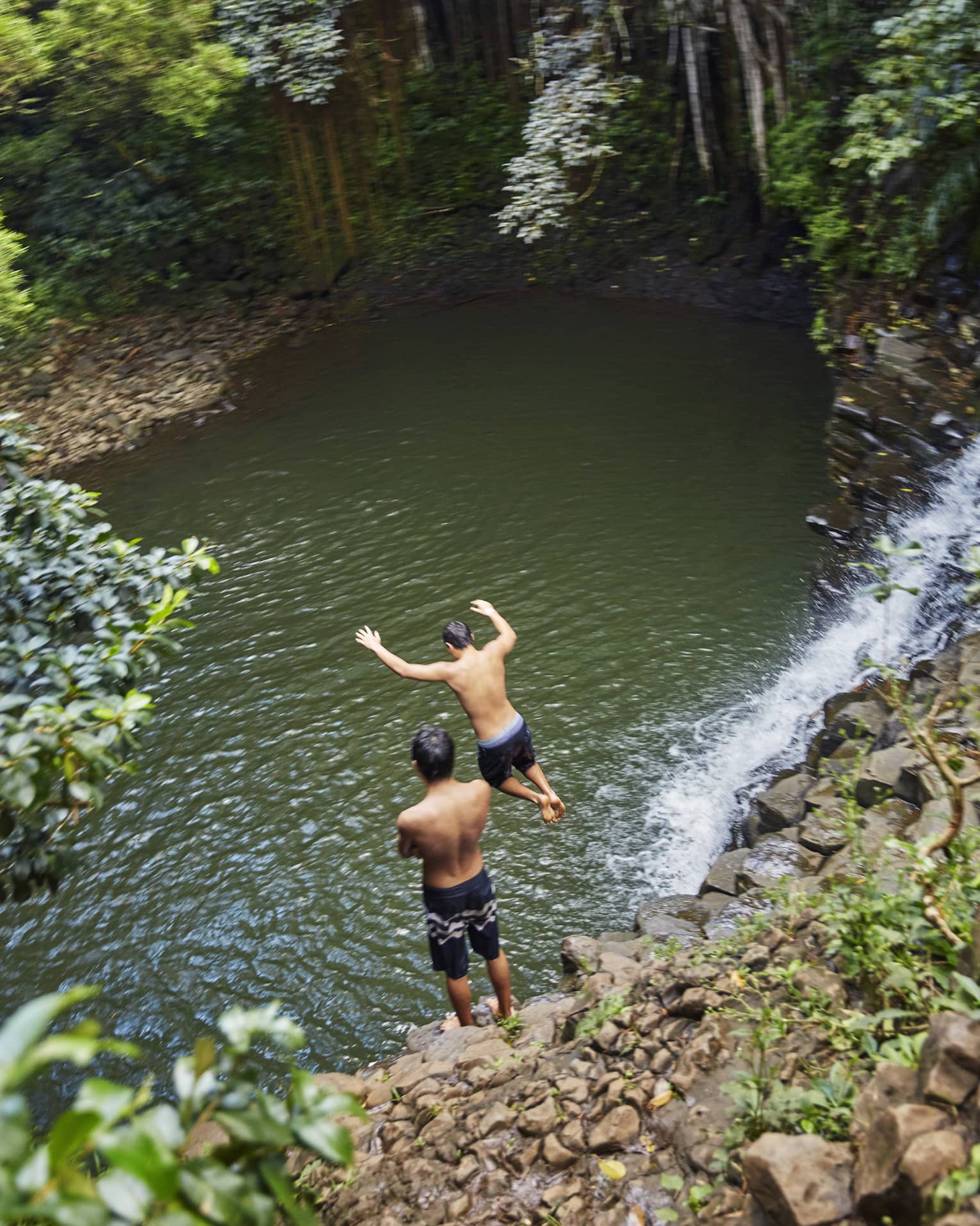 Two people leap from rocks to water below waterfall on the road to Hana