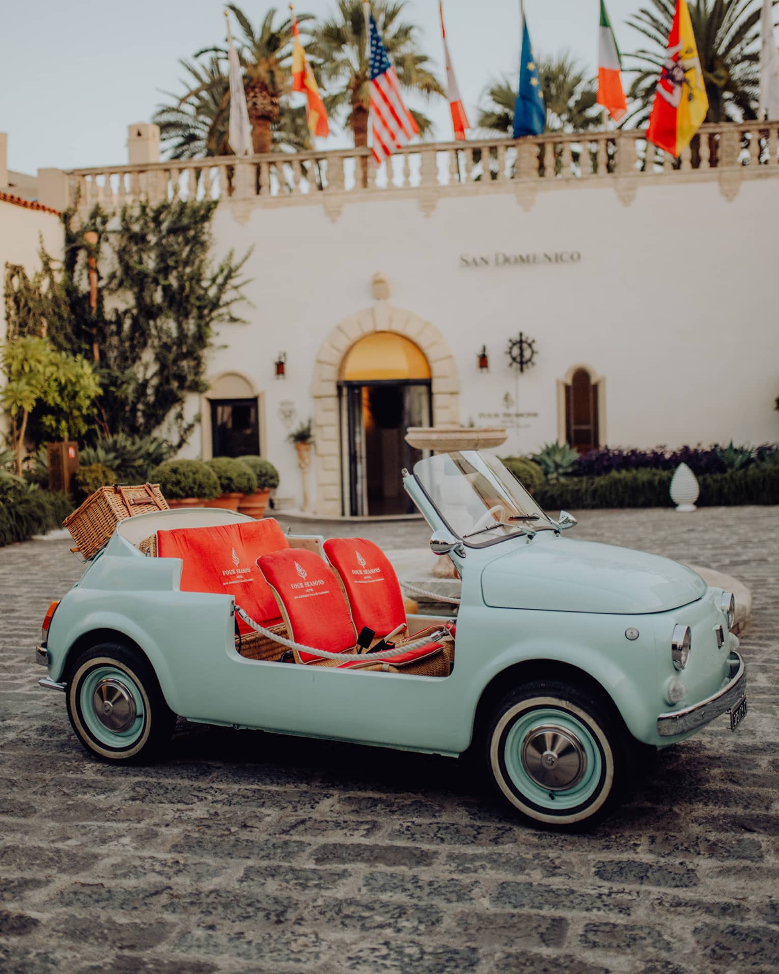 Vintage teal convertible Fiat 500 with bright coral seats parked on a cobblestone driveway in front of San Domenico Palace.  