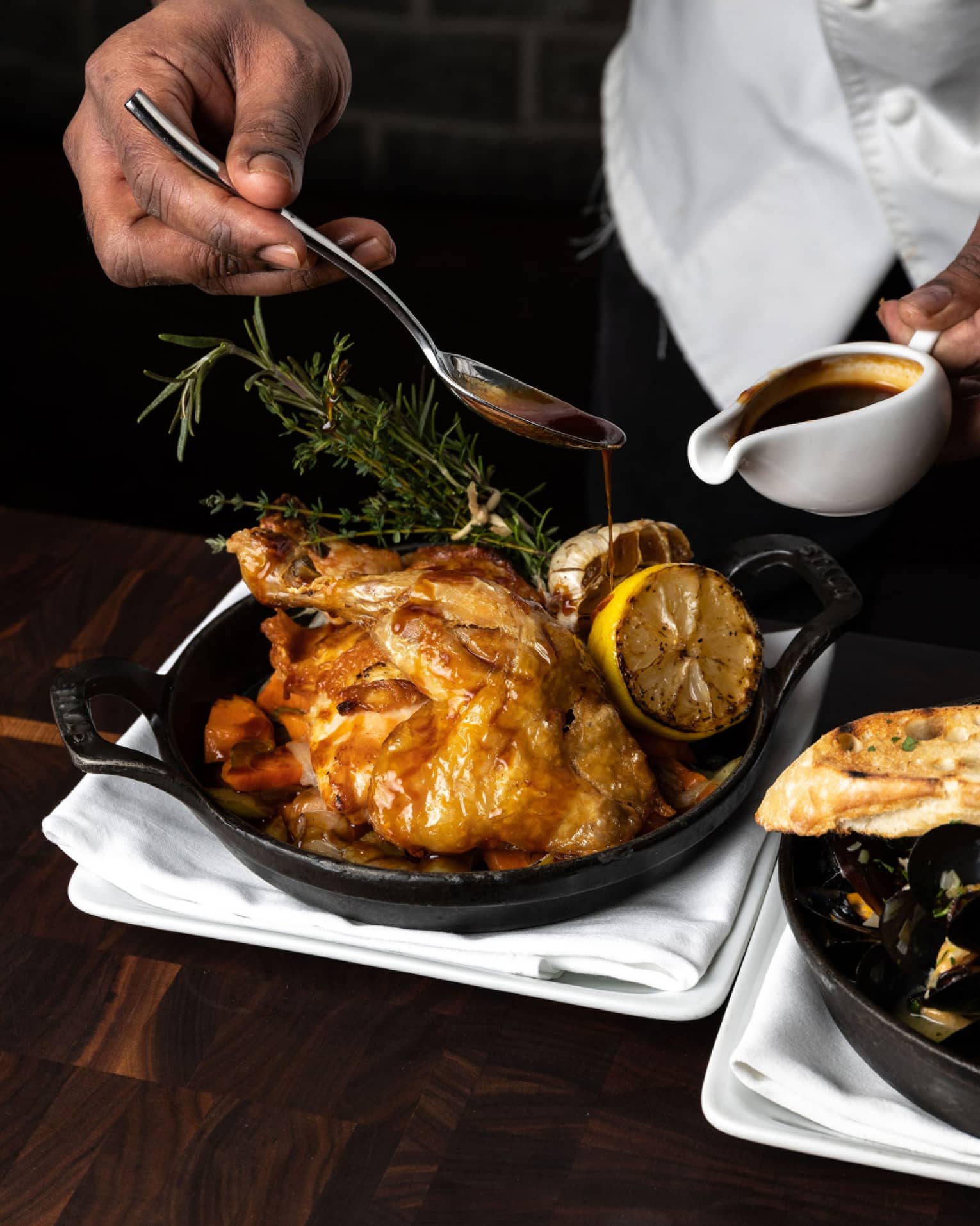 Chef's hands basting a full roasted chicken in an iron skillet, garnished with greenery and roasted lemons