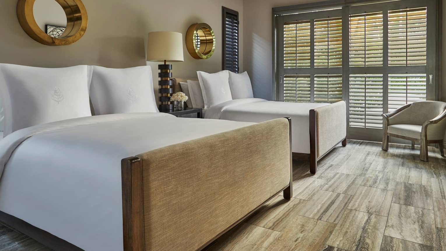 Luxury resort bedroom with two beds, white bedding, decorative mirrors, a nightstand with a lamp and shuttered windows letting in natural light