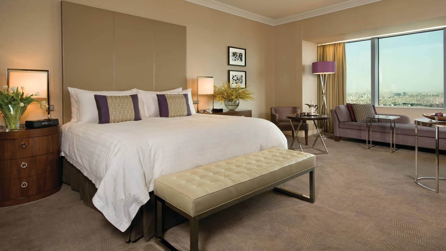 Deluxe hotel room bed with tall padded headboard and leather bench, beige carpet, long chaise at window
