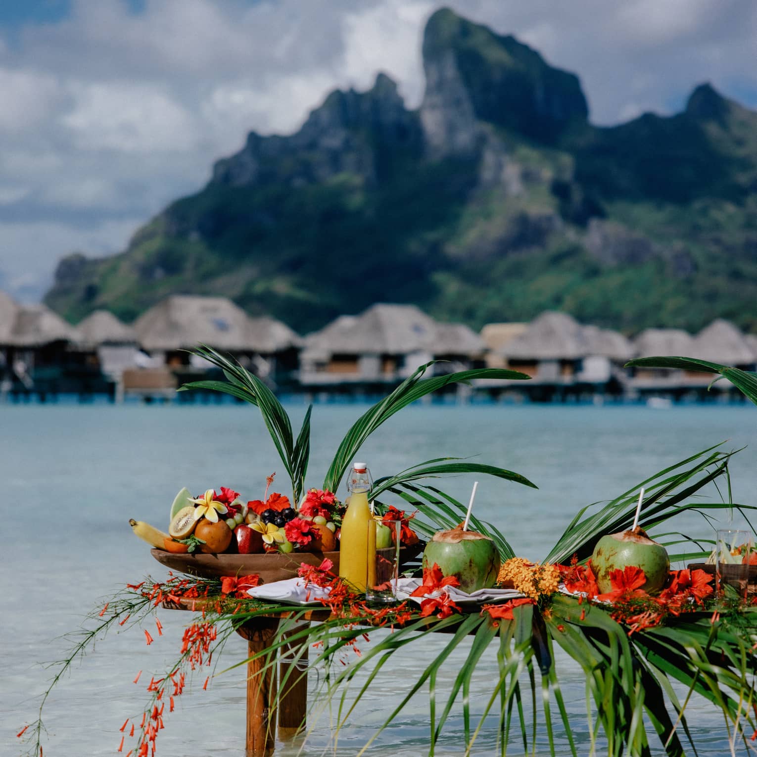 Small wooden table with legs submerged in clear water, topped with assorted fruit and coconuts with straws, mountain and overwater pavilions in background
