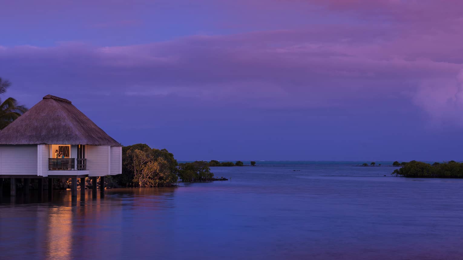 Spa staff massaging woman's head on deck of small white villa over water at night
